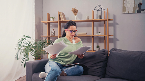 Confused student in eyeglasses looking at documents on couch