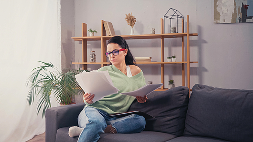 Concentrated student in eyeglasses looking at documents on couch in living room