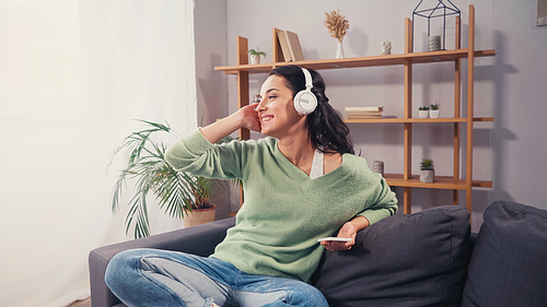 Cheerful woman in headphones holding smartphone on couch