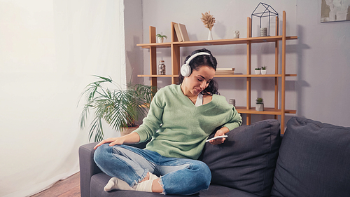Smiling woman in wireless headphones using smartphone on couch
