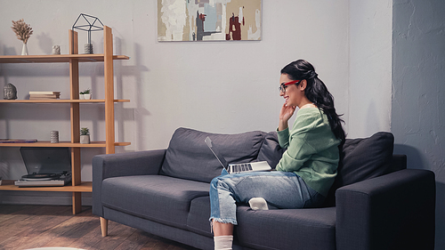 Side view of positive woman in eyeglasses looking at laptop on couch