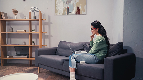 Side view of pretty teleworker in eyeglasses using laptop on couch