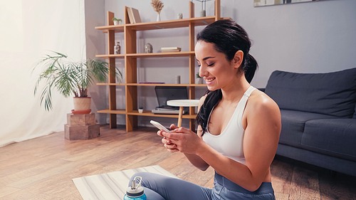 Smiling sportswoman using smartphone near sports bottle and fitness mat at home