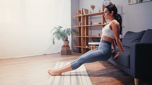 Side view of athletic woman working out near couch and fitness mat at home