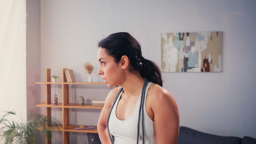 Side view of tired sportswoman with jump rope standing in living room