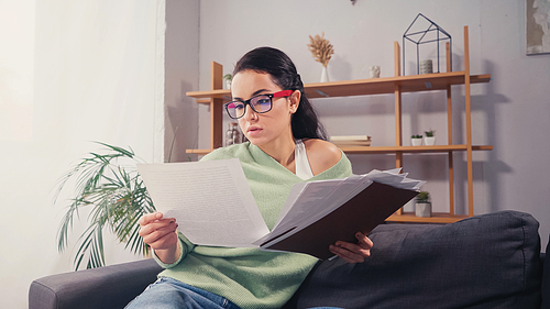 Young student in eyeglasses holding papers and paper folder on couch