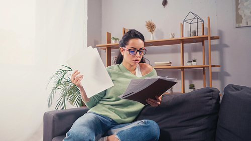 Young brunette student in eyeglasses looking at documents in living room