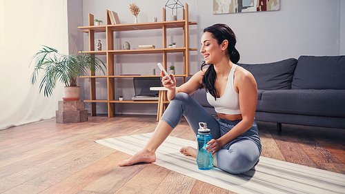 Side view of cheerful sportswoman using mobile phone on fitness mat near sports bottle at home
