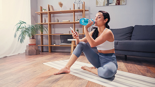 Sportswoman drinking water and using cellphone on fitness mat in living room