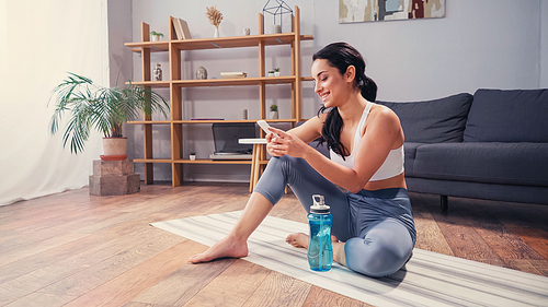 Positive woman in sportswear using smartphone near sports bottle at home