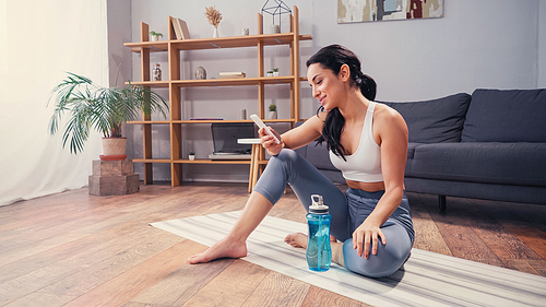 Side view of sportswoman using cellphone while resting near sports bottle on fitness mat