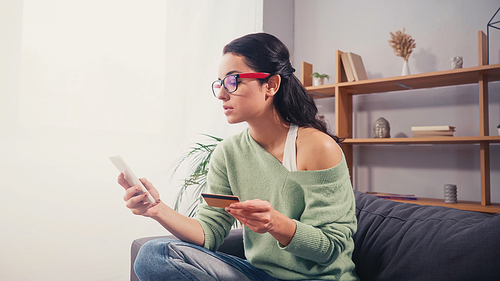 Young woman in eyeglasses using smartphone and credit card during online shopping at home