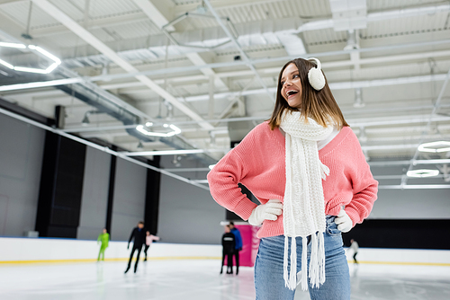 excited woman in ear muffs and pink sweater posing with hands on hips on ice rink