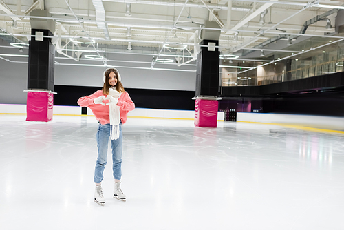 full length of happy woman in white scarf and ear muffs showing heart with hands while skating on ice rink