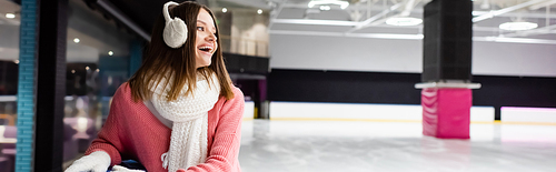 excited young woman in ear muffs and scarf laughing on ice rink, banner
