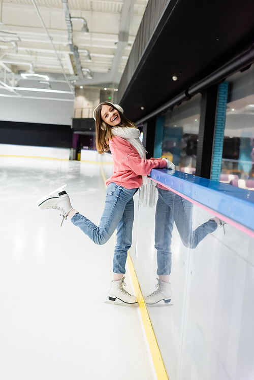full length of happy woman in sweater, white scarf and ear muffs posing on ice rink