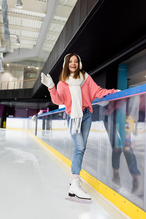 full length of happy young woman in winter outfit skating on frozen ice rink and waving hand