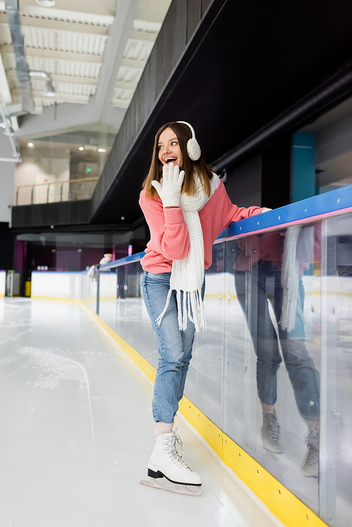 full length of excited brunette woman in gloves and ear muffs on ice rink