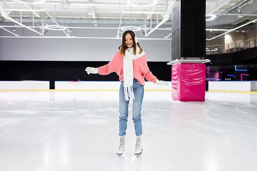 full length of happy woman in knitted sweater, ear muffs and winter outfit skating on ice rink