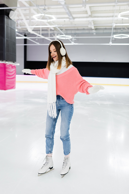 happy young woman in white ear muffs and knitted scarf skating with outstretched hands on ice rink