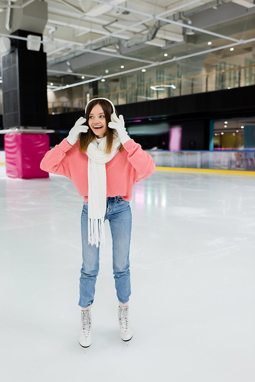 full length of cheerful young woman in white ear muffs and winter outfit skating on frozen ice rink