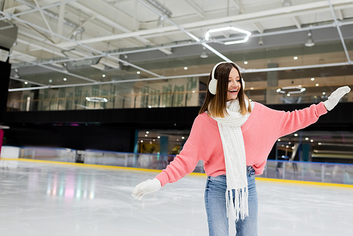 excited woman in white ear muffs and pink sweater skating with outstretched hands on ice rink