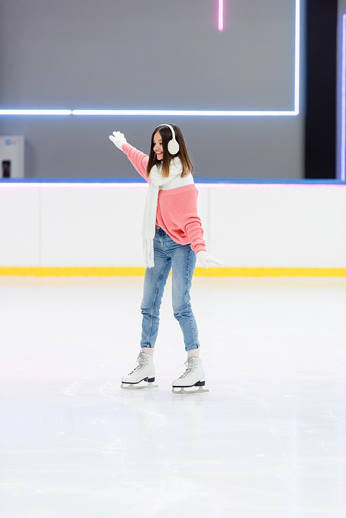 full length of smiling young woman in ear muffs and scarf skating with outstretched hands on ice rink