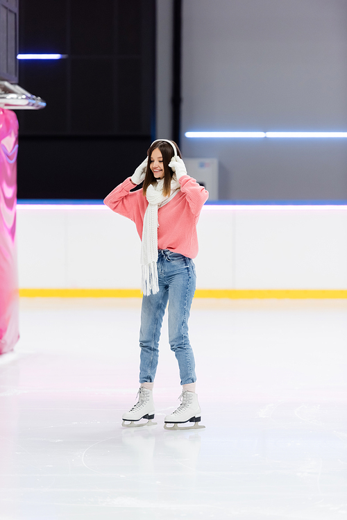 full length of cheerful woman in white scarf adjusting ear muffs and skating on ice rink
