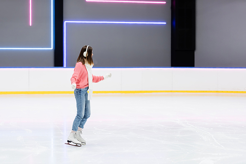 full length of young woman in knitted sweater, ear muffs and scarf skating on ice rink