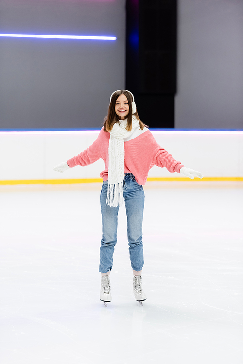 full length of cheerful woman in white ear muffs and scarf skating with outstretched hands on ice rink