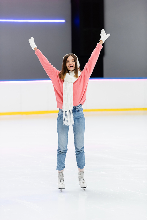 full length of happy woman in knitted sweater, ear muffs and winter outfit skating with raised hands on ice rink