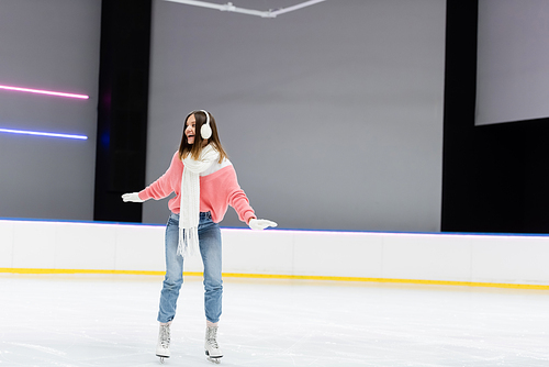 full length of happy woman in ear muffs and winter outfit skating on frozen ice rink