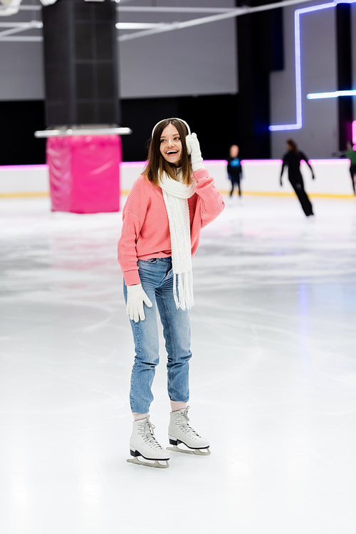 full length of happy woman in sweater, white scarf and ear muffs skating on ice rink