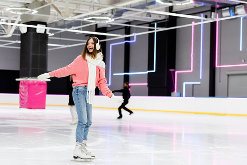 full length of cheerful young woman in winter outfit skating with outstretched hands on ice rink