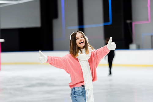 amazed young woman in pink sweater showing thumbs up on ice rink