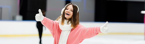 happy young woman in ear muffs and scarf showing thumbs up on ice rink, banner