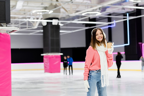 happy woman in ear muffs and pink sweater holding paper cup on ice rink