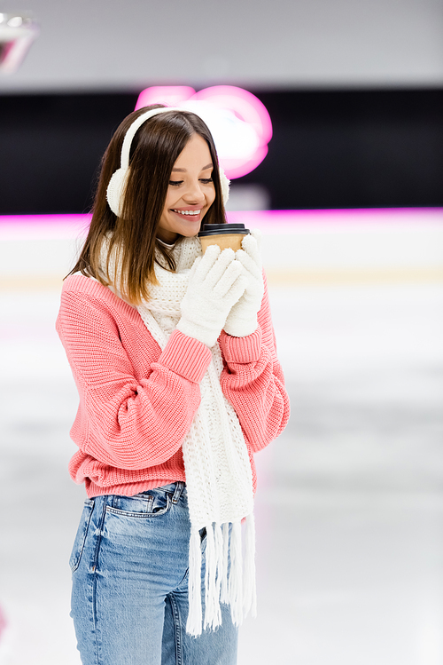 happy woman in white gloves and ear muffs holding paper cup on ice rink