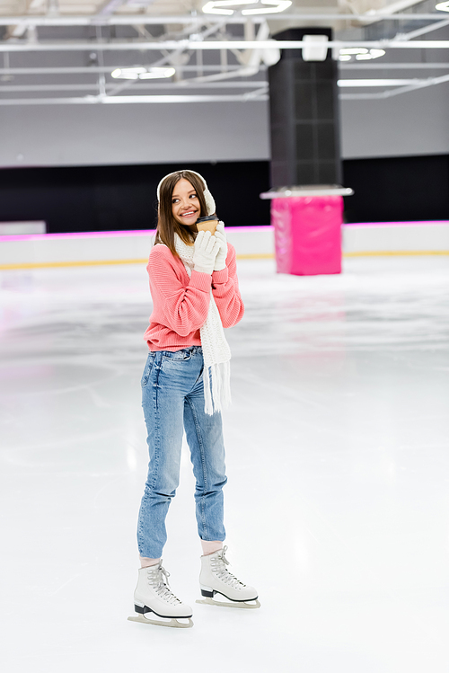 full length of happy woman in knitted sweater, ear muffs and winter outfit skating with paper cup on ice rink