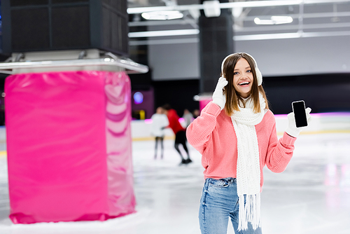 amazed woman in white ear muffs holding smartphone with blank screen on ice rink