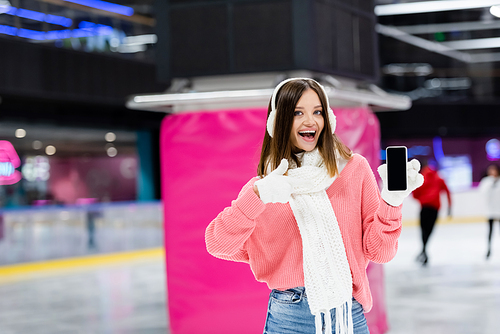 amazed woman in white ear muffs holding smartphone with blank screen and showing thumb up on ice rink