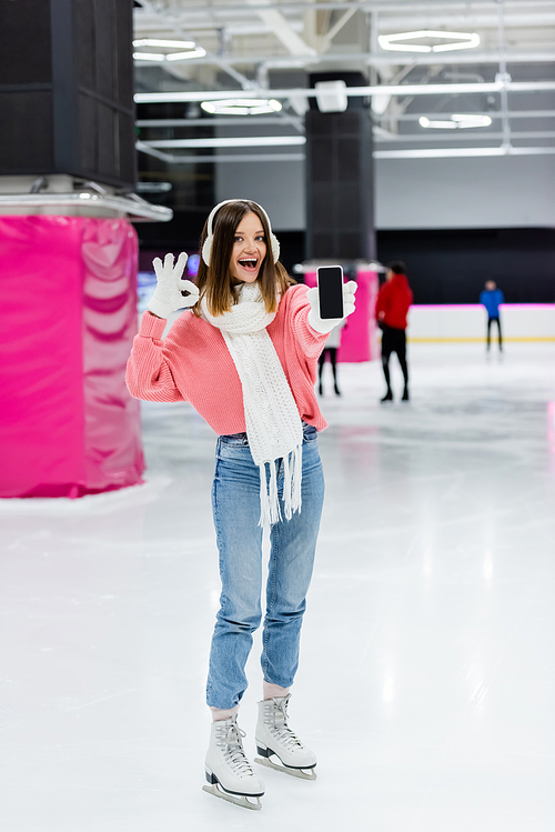 excited woman in gloves and ear muffs showing ok sign while holding smartphone with blank screen on ice rink