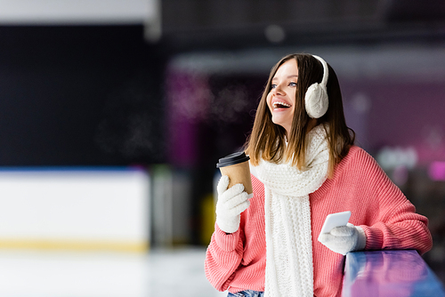 excited woman in ear muffs holding paper cup and smartphone on ice rink