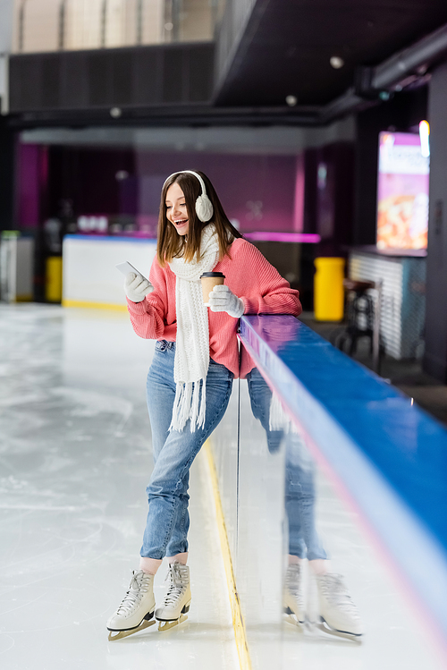full length of young woman with paper cup and smartphone standing on ice rink