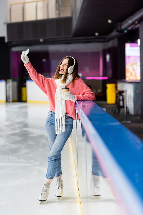 positive young woman with paper cup taking selfie on ice rink