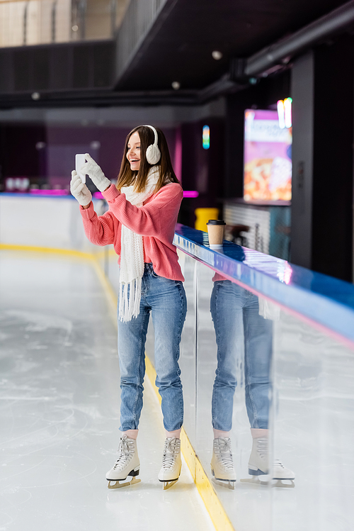 full length of brunette woman in gloves and ear muffs taking photo on ice rink
