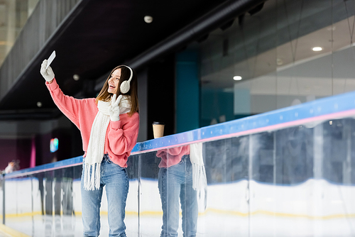 positive young woman taking selfie while showing peace sign near paper cup on ice rink