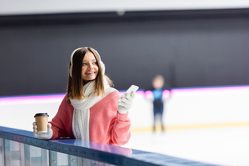 pleased woman in white ear muffs and pink sweater holding paper cup and cellphone on ice rink