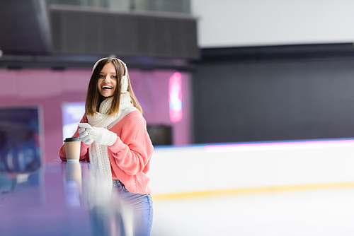 excited woman in ear muffs holding smartphone near paper cup on ice rink