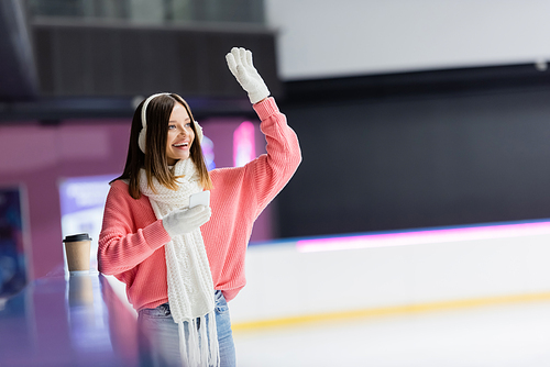 happy woman in ear muffs holding smartphone and waving hand on ice rink